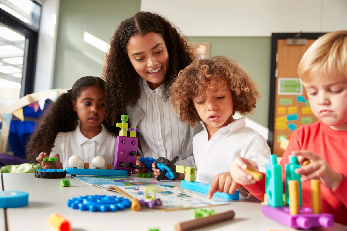 Female teacher sitting at table in play room with three kindergartne children constructing, selective focus Female teacher sitting at table in play room with three kindergartne children constructing, selective focus