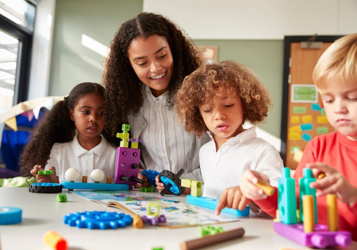 Female teacher sitting at table in play room with three kindergartne children constructing, selective focus