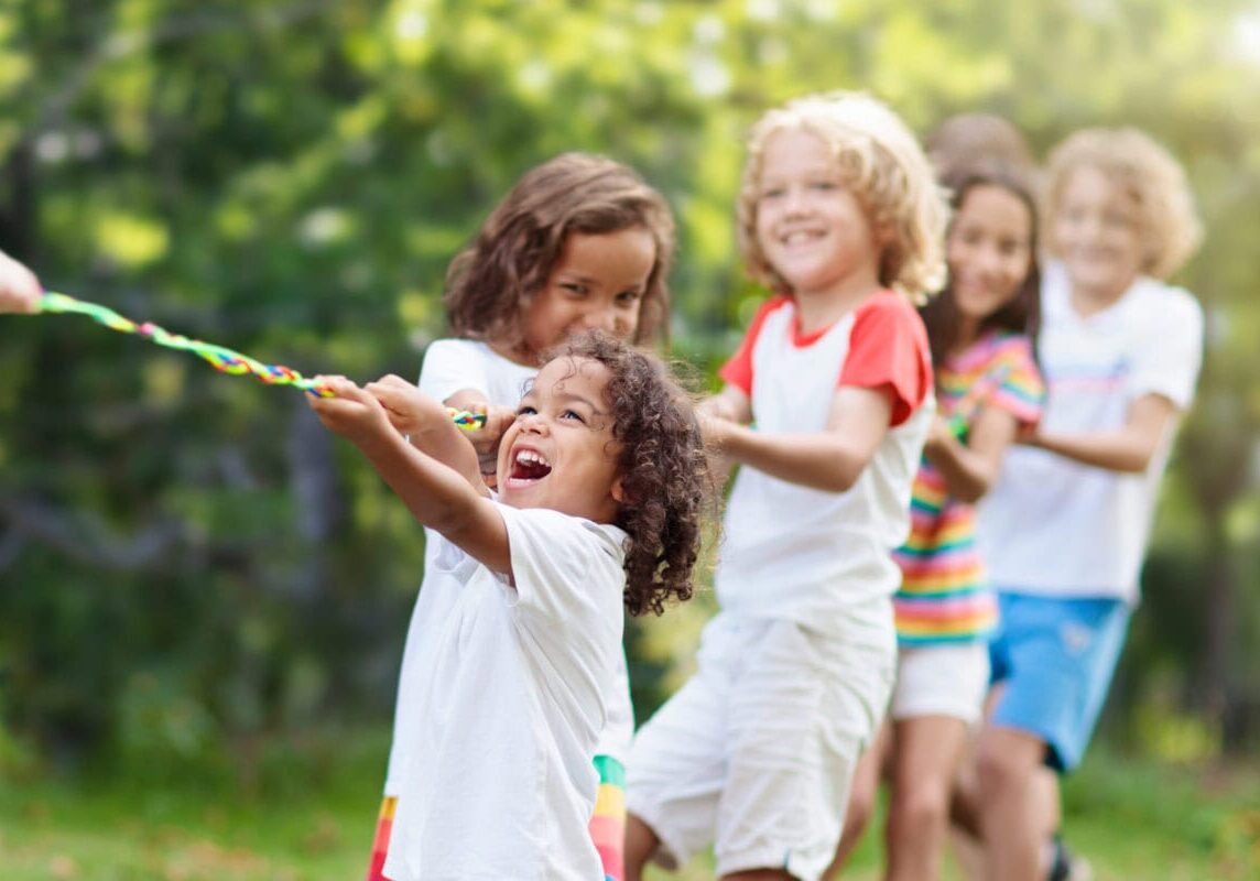 Kids play tug of war in sunny park. Summer outdoor fun activity. Group of mixed race children pull rope in school sports day. Healthy outdoor game for little boy and girl.